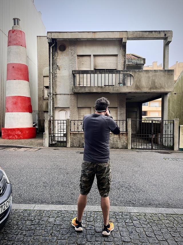 Un photographe se tient sur un trottoir pavé, face à une maison en mauvais état, avec des volets fermés et des murs usés. À gauche de la maison, il y a une grande structure cônique, peinte de bandes rouges et blanches, ressemblant à un phare.