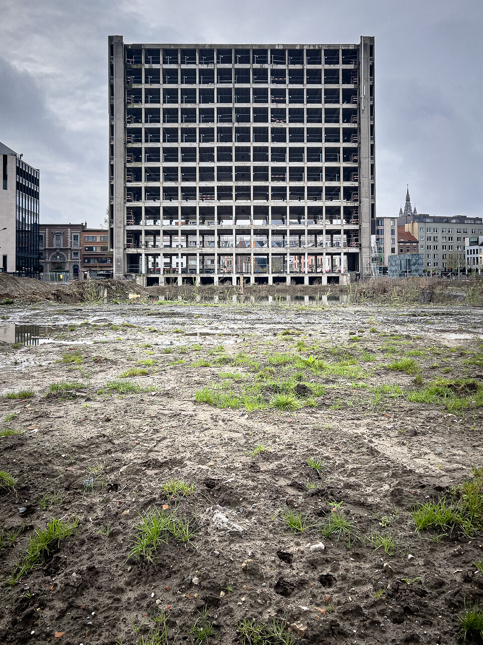 Ancien bâtiment RTT/Belgacom en cours de démolition, avec une structure en béton visible et de nombreuses ouvertures rectangulaire, sol boueux, ciel nuageux, ambiance grise et morose.
