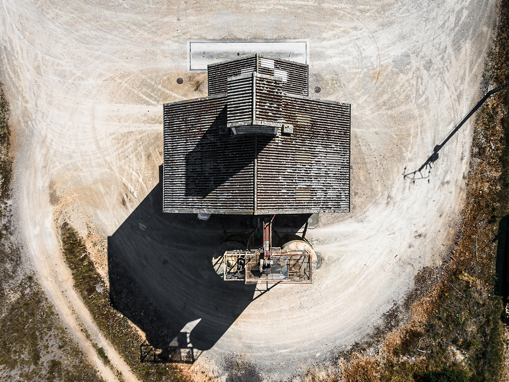 Vue aérienne d'un silo situé dans les environs de Dijon, avec un toit nervuré et patiné. Le bâtiment est entouré de terre battue et de gravier, avec des traces de véhicules formant des courbes autour de la structure. L'ombre portée du bâtiment est longue et sombre, indiquant un soleil bas. Au premier plan, on distingue quelques éléments de machinerie ou de tuyauterie juste à côté de la structure centrale.