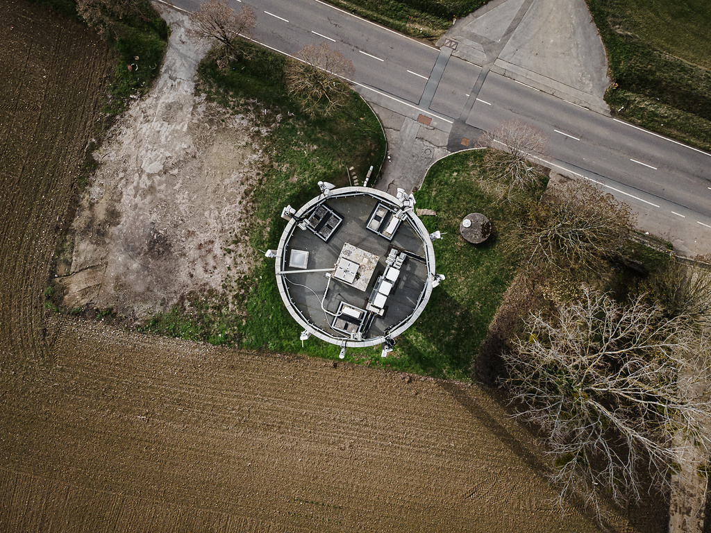 Vue aérienne du château d'eau de forme circulaire à Elvange, montrant le toit technique. La structure est située le long d'une route, bordée par des champs agricoles et des arbres partiellement dénudés.