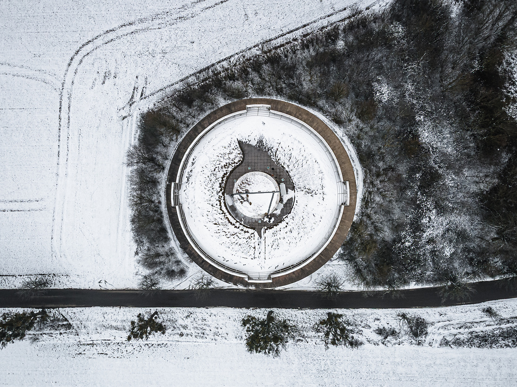 Vue aérienne du château d'eau de Remerschen recouvert de neige en hiver. La structure circulaire avec son design géométrique est bordée par une route, des champs enneigés et une zone forestière.