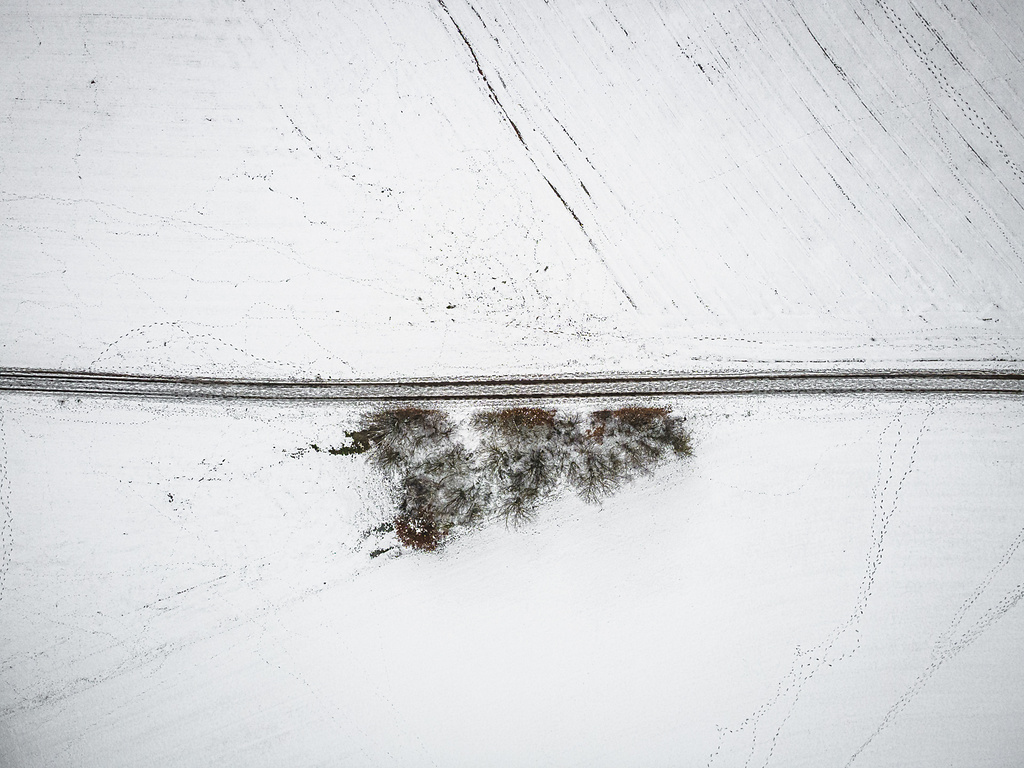 Vue aérienne d'une route de campagne sombre et horizontale, coupant des champs agricoles enneigés en hiver. Un petit bosquet d'arbres dénudés se trouve près de la route.