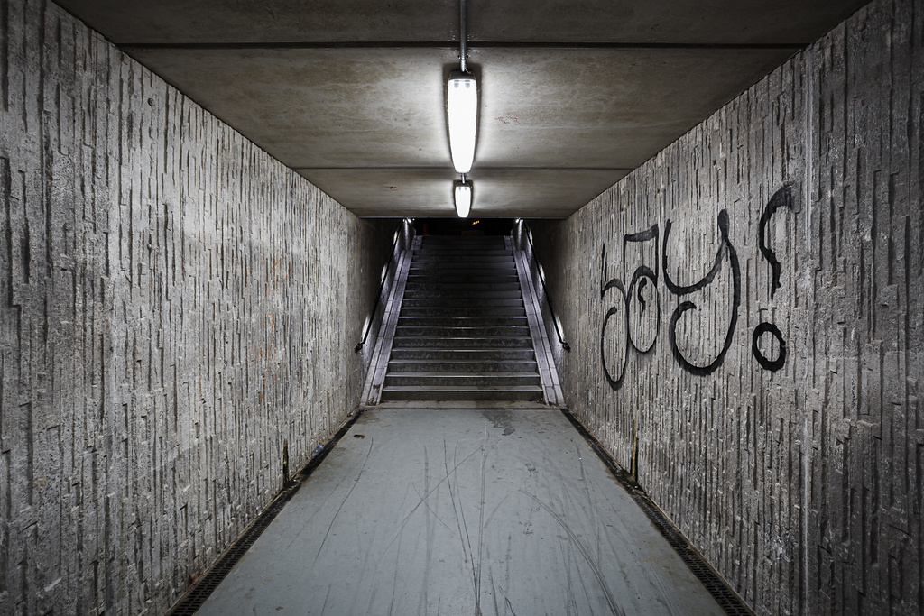 Tunnel en béton avec des murs texturés couverts d'un graffiti. Au bout, un escalier, le tout éclairé par des néons, créant une atmosphère mystérieuse.