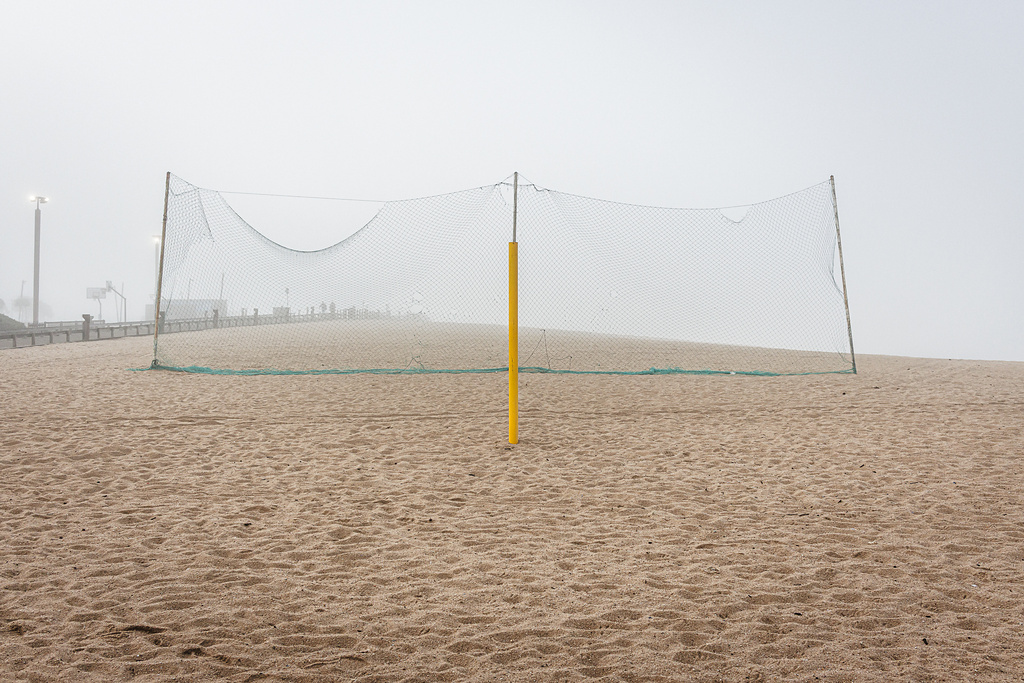 Plan large et symétrique d'un terrain de beach-volley déserté, sous une lumière diffuse due au brouillard. Au centre, un filet vert séparant le terrain de la plage et un poteaux de couleur jaune à l'avant plan.