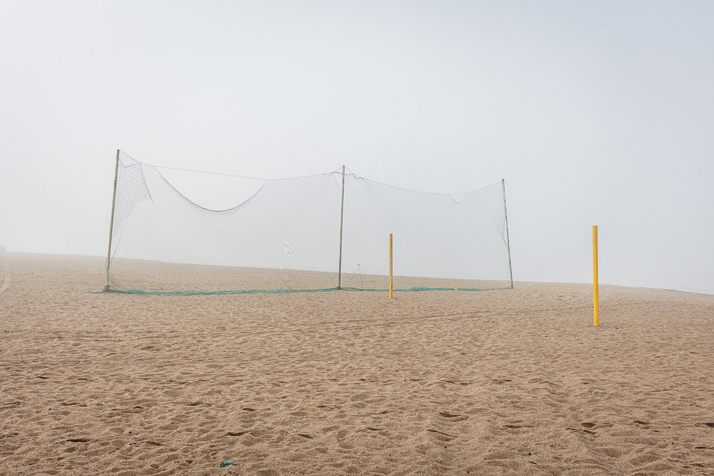 Plan large et minimaliste d'un terrain de beach-volley déserté, sous une lumière diffuse due au brouillard. Au centre, un filet vert est tendu pour séparer le terrain de la plage. Deux poteaux de couleur jaune, sans filet, se dressent à droite dans le sable.