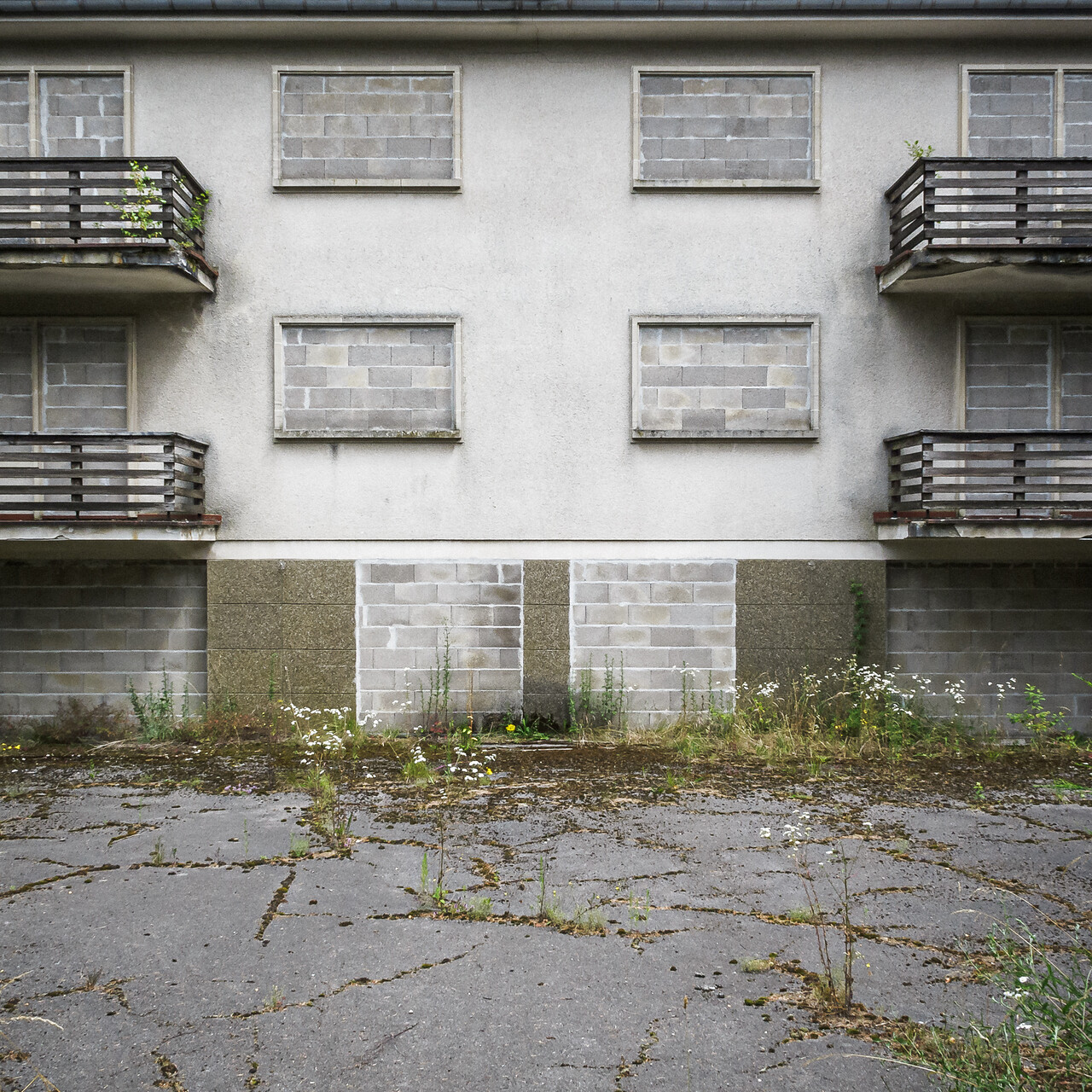 Photog rapprochée de la façade d'une résidence abandonnée à Senningerberg. Les fenêtres du rez-de-chaussée et du premier étage sont entièrement occultées par des parpaings. Au premier plan, de la végétation sauvage pousse à travers les fissures de l'asphalte.