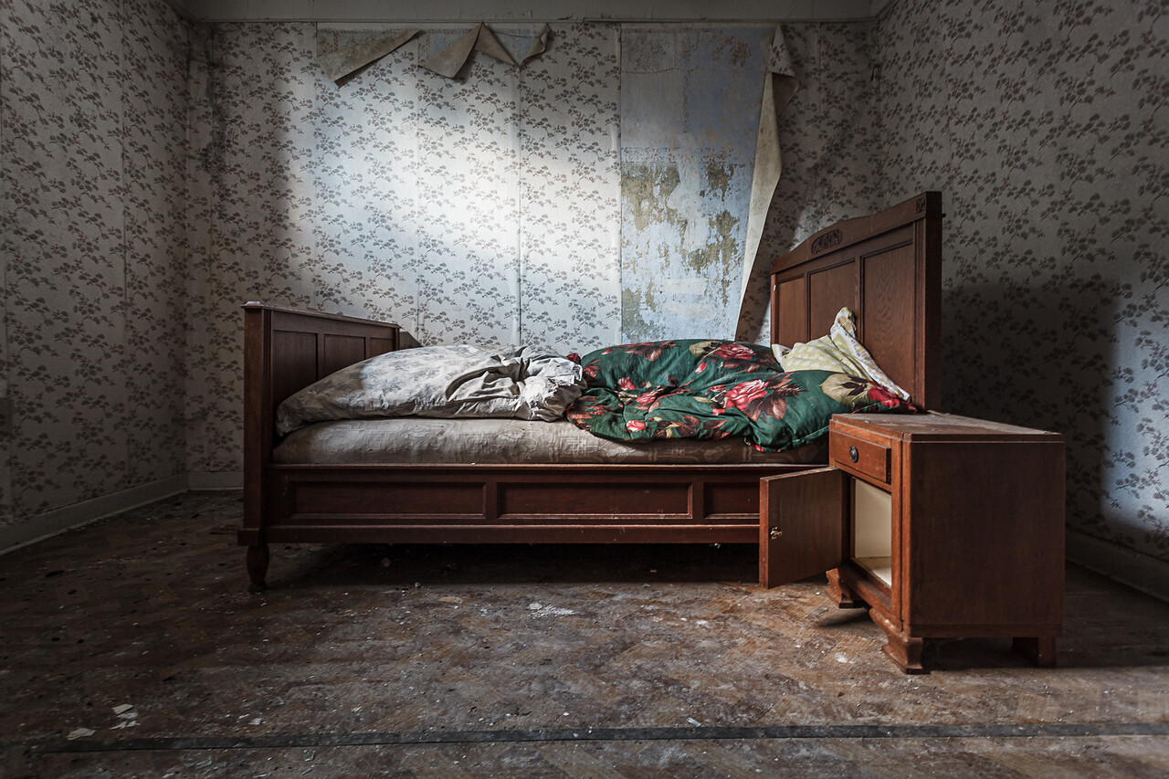 Grand angle d'un vieux lit et sa table de chevet dans une chambre décorée de papier peint à motifs...