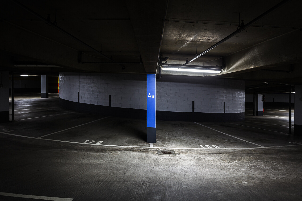 Parking souterrain sombre à Esch-sur-Alzette, Luxembourg, avec un pilier bleu portant la référence '4B' au centre, des marquages au sol blancs numérotés '179' et '180', le sol en béton incurvées et un éclairage aux néons.