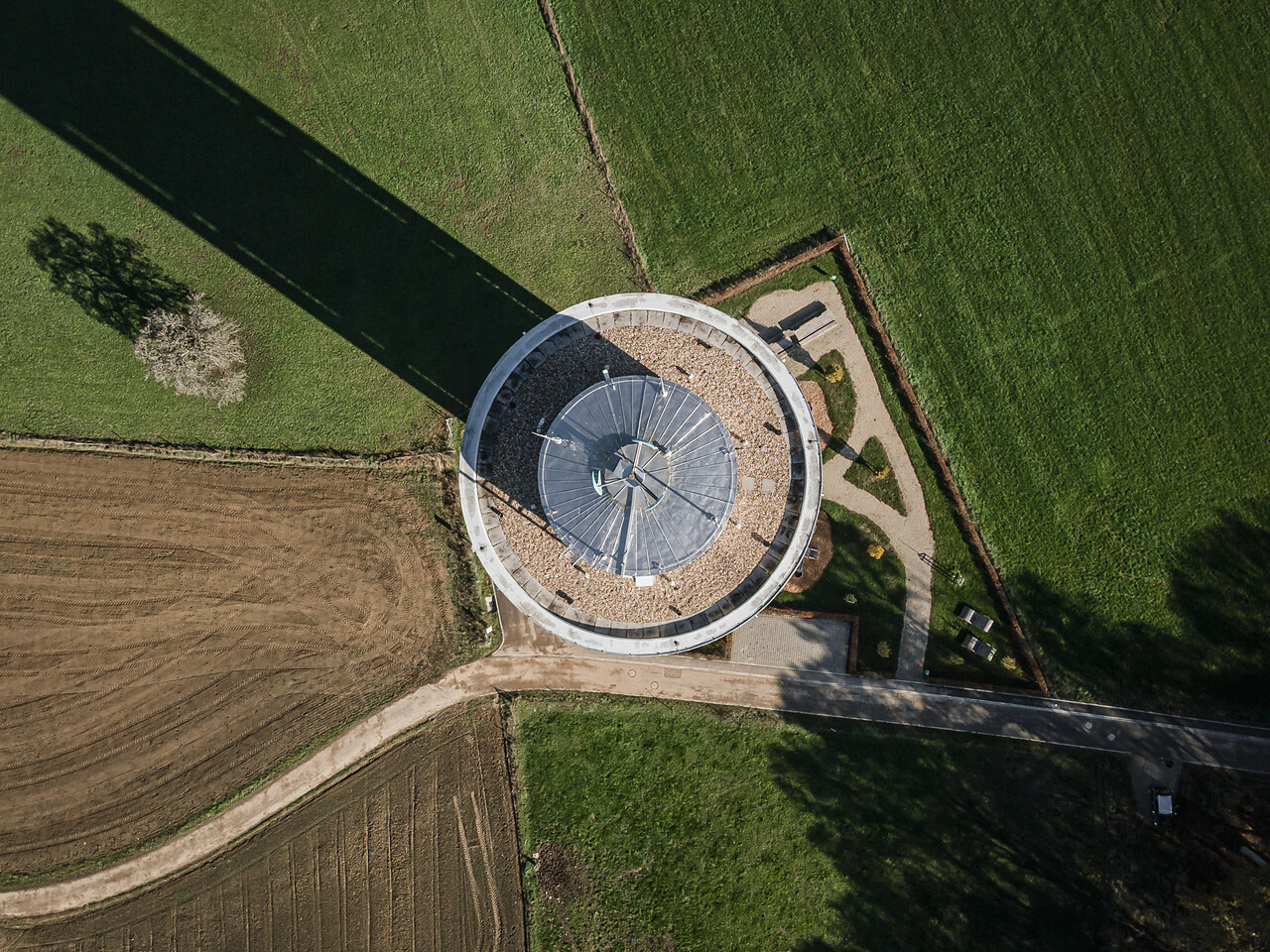 Vue aérienne d'un château d'eau avec un toit gris, entouré de champs agricoles verts et bruns. Une longue ombre noire et nette s'étend à l'arrière-plan, suggérant une structure élancée ou un mât.