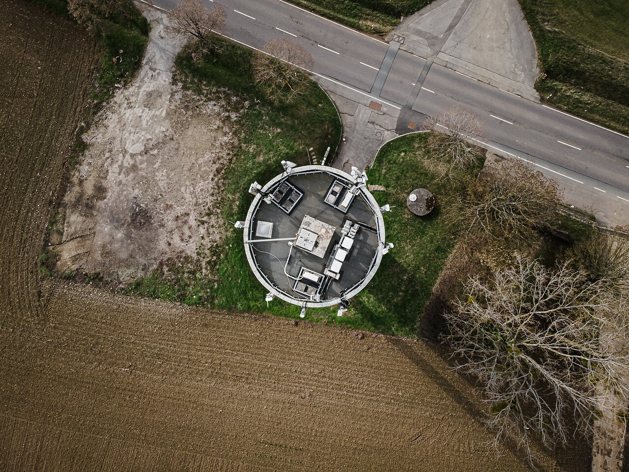 Vue aérienne du château d'eau de forme circulaire à Elvange, montrant le toit technique. La structure est située le long d'une route, bordée par des champs agricoles et des arbres partiellement dénudés.