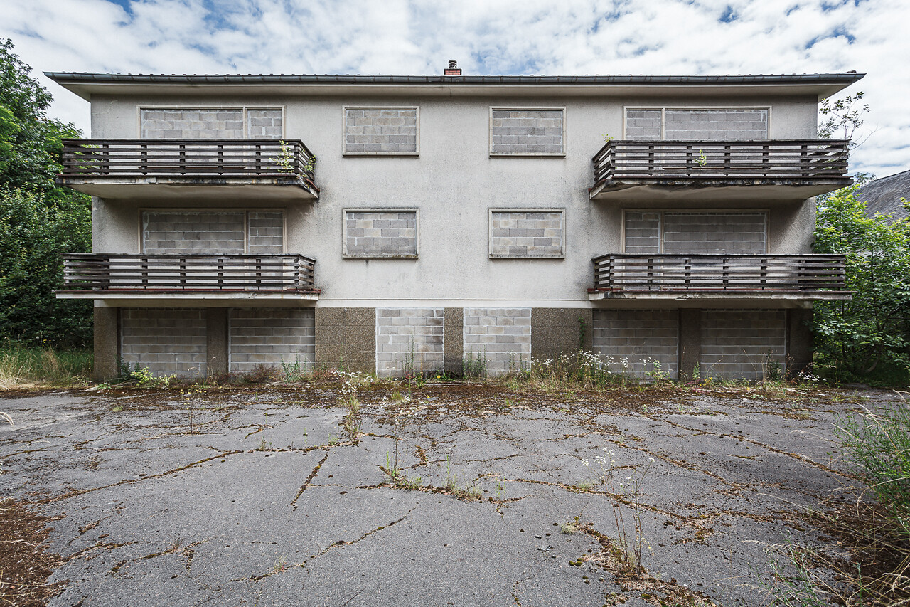 Vue d'ensemble symétrique du bâtiment abandonné au 30-32 route de Trèves. La structure de trois étages, murée à tous les niveaux, se dresse sous un ciel partiellement nuageux.
