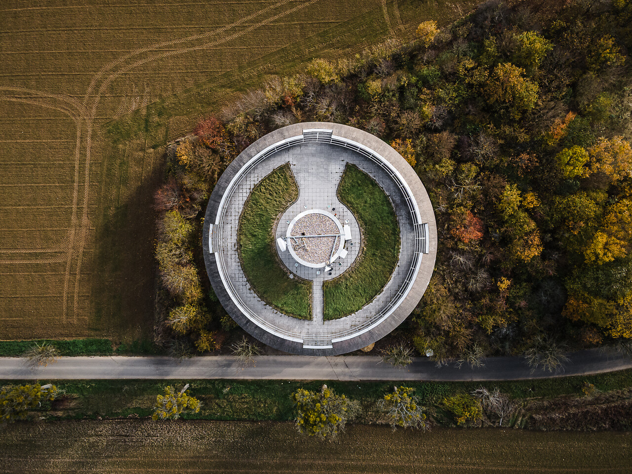 Vue aérienne du château d'eau de Remerschen, remarquable par sa plateforme au sommet composée de béton et pelouse. La structure est encadrée par une route, des champs labourés et une forêt aux couleurs d'automne.