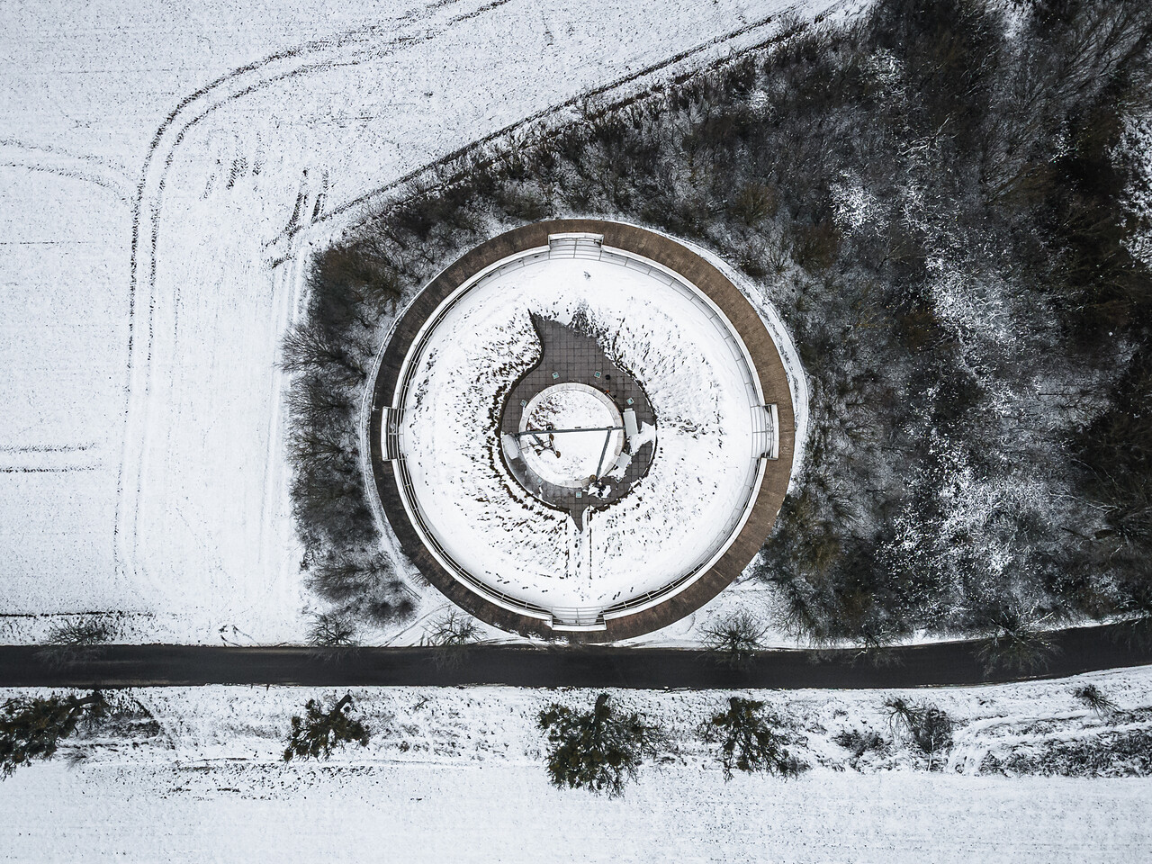 Vue aérienne du château d'eau de Remerschen recouvert de neige en hiver. La structure circulaire avec son design géométrique est bordée par une route, des champs enneigés et une zone forestière.