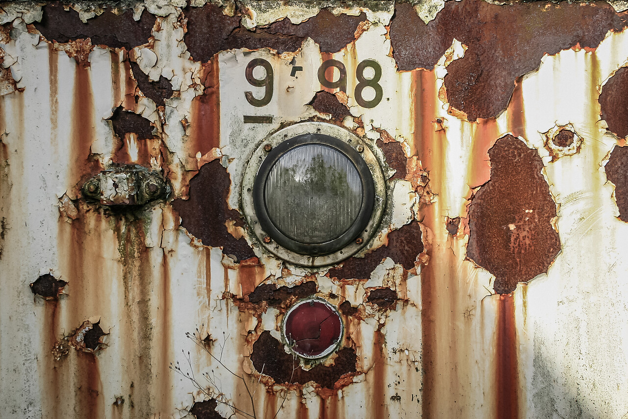 Face avant rouillée et écaillée d'un tram, deux phares circulaires (l'un transparent et l'autre rouge), et l'inscription '9?98' en haut.