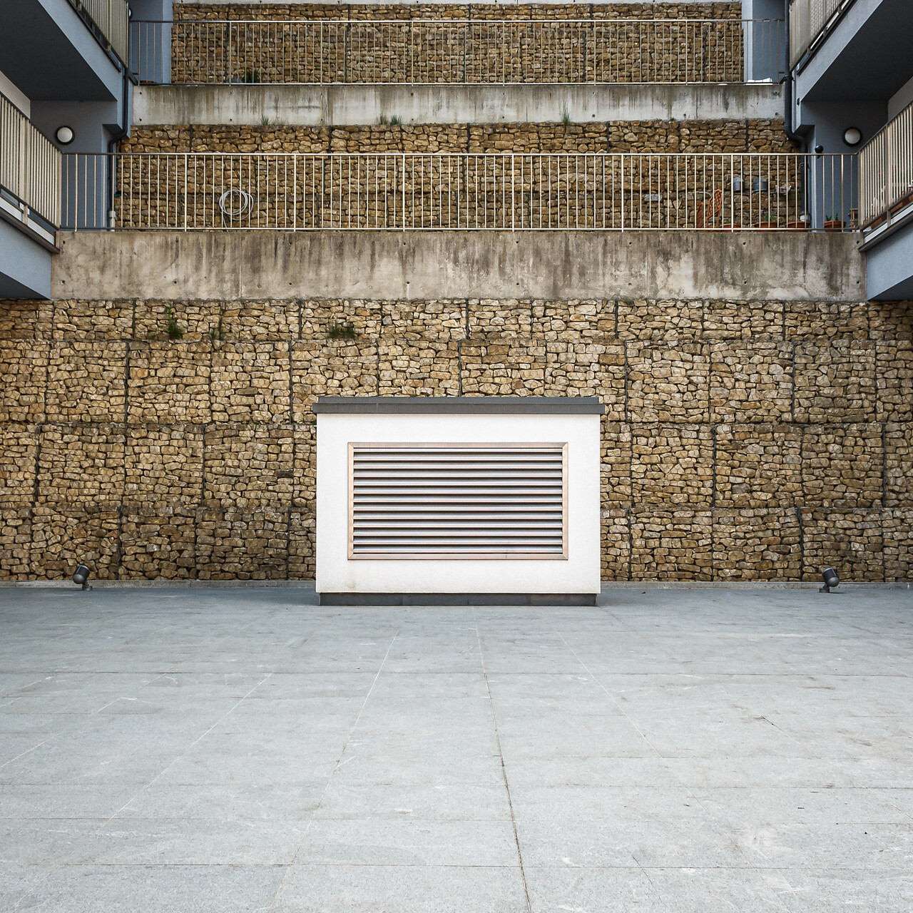 Cour moderne pavée de dalles grises, avec au centre une bouche d’aération blanche à lames horizontales, adossée à un mur en pierres empilées dans une cage métallique; balcons latéraux avec rampes métalliques et appliques murales, architecture mêlant matériaux bruts et lignes épurées.