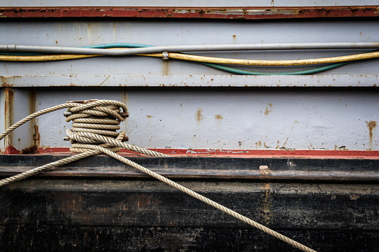 Un cordage enroulé autour d'un taquet sur une péniche, avec des tuyaux et des câbles fixés sur la paroi métallique rouillée en arrière-plan.