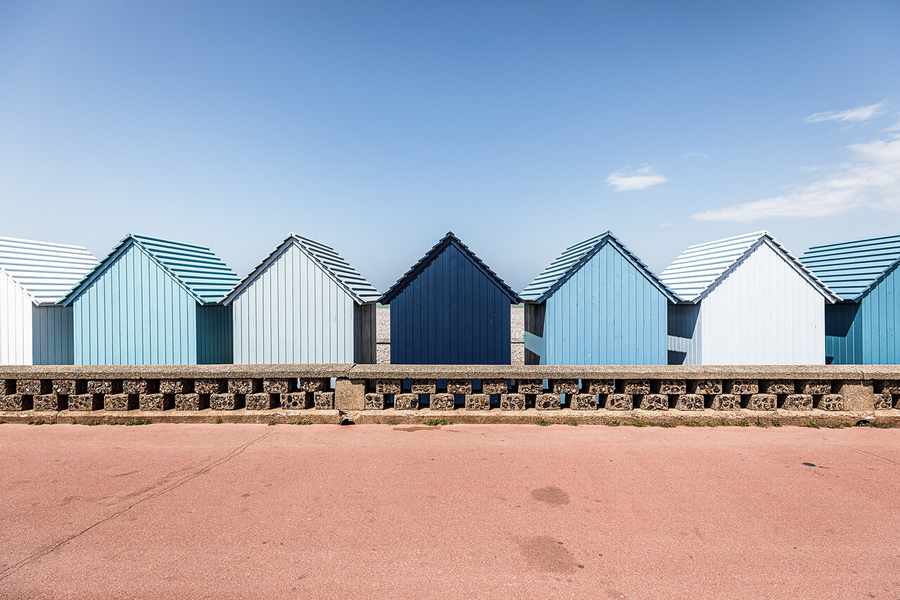 Une rangée de cabines de plage en bois aux dégradés de bleu, alignées derrière un muret en pierre sur le front de mer de Dieppe. Au centre, une cabine bleu marine se détache parmi d'autres aux tons bleu ciel et blanc cassé. Le premier plan montre une promenade rose saumoné sous un ciel bleu clair et dégagé.