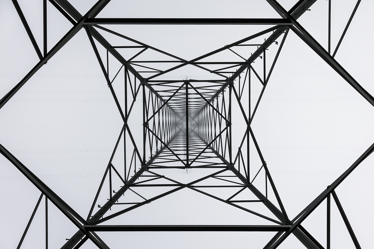 Vue en contre-plongée d'un pylône électrique, dont la structure métallique complexe forme une cage géométrique avec des lignes électriques horizontales.