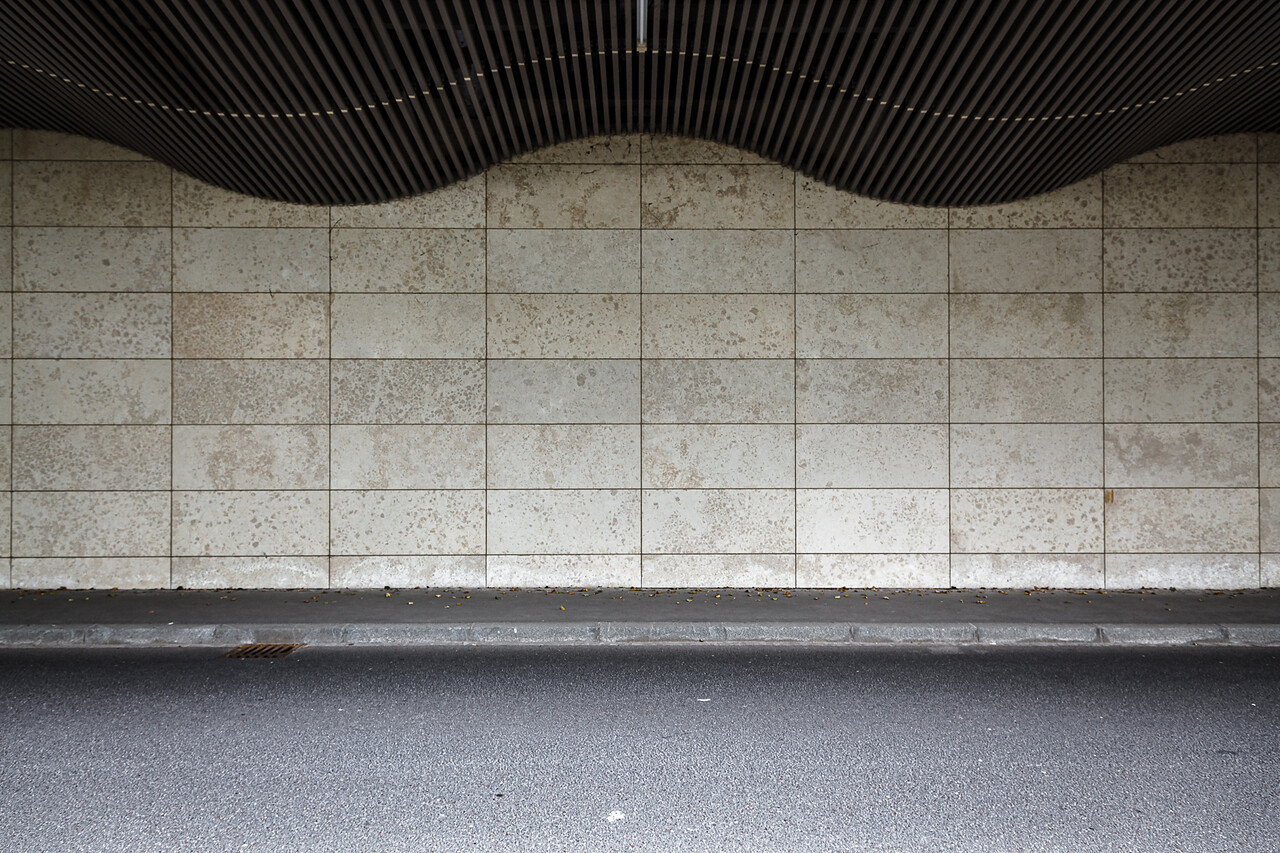 Pan de un mur carrelé et une structure en surplomb. Le mur est constitué de carreaux rectangulaires avec une apparence tachetée et légèrement vieillie. Au-dessus, une marquise ondulée composée de lattes verticales espacées crée un motif distinctif. Devant le mur se trouvent un trottoir et une portion de route asphaltée.
