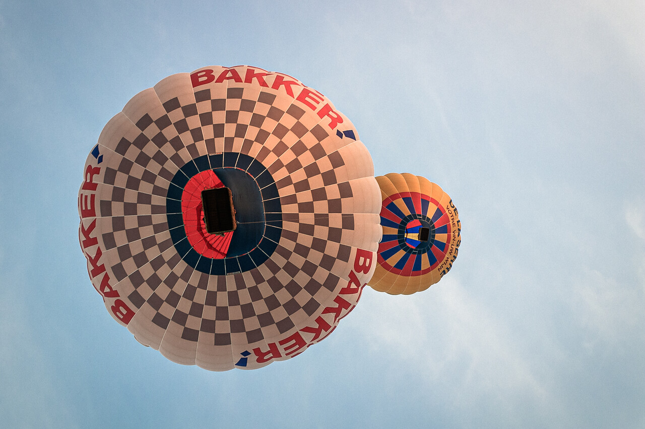 Deux montgolfières vues d'en dessous à Baisy-Thy en Belgique. La montgolfière principale est à damier gris et blanc avec l'inscription 'BAKKER!' répétée sur le bord, et une deuxième montgolfière plus petite est visible en arrière-plan avec des motifs circulaires rouge, orange et bleus.