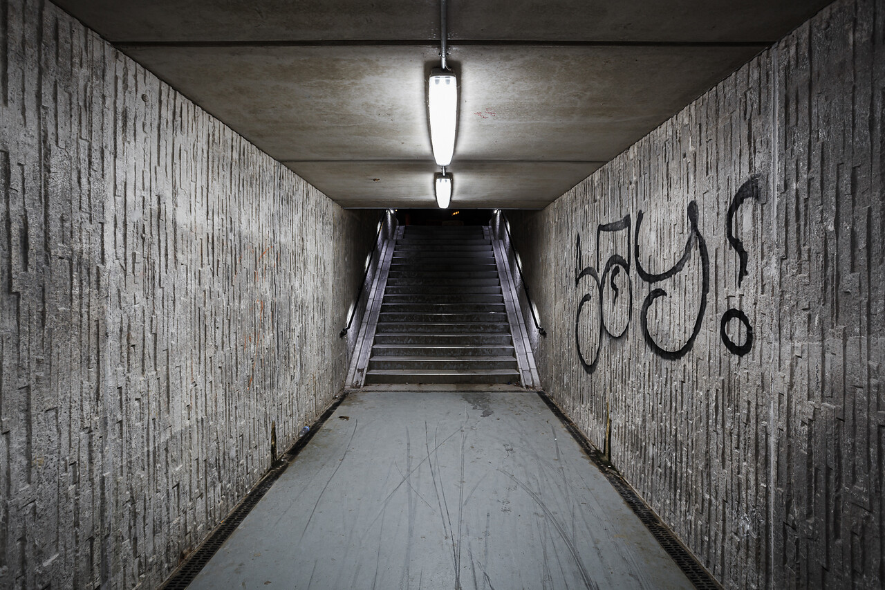 Tunnel en béton avec des murs texturés couverts d'un graffiti. Au bout, un escalier, le tout éclairé par des néons, créant une atmosphère mystérieuse.