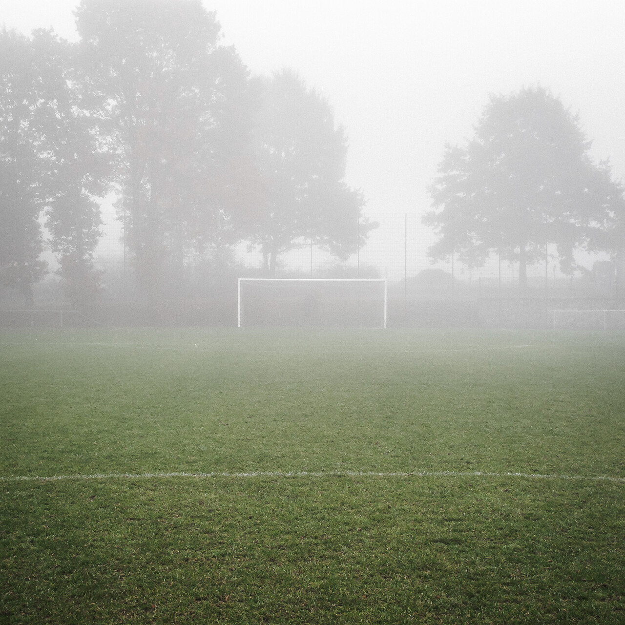 La photo représente un terrain de football par temps de brouillard. Le gazon est vert, un but blanc se trouve au centre et des arbres en arrière-plan sont partiellement cachés par le brouillard.