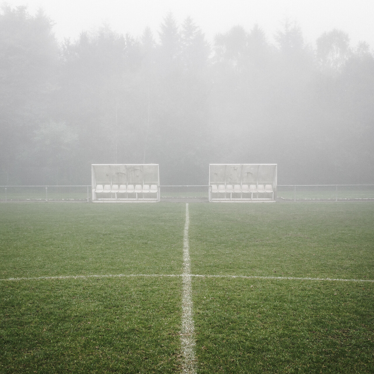 L'image montre un terrain de football embrumé, avec deux bancs d'équipe vides placés de part et d'autre de la ligne médiane. Le terrain est recouvert d'herbe verte, et une ligne blanche marquant le centre divise l'image verticalement. À l'arrière-plan, une forêt dense d'arbres hauts est partiellement masquée par le brouillard.