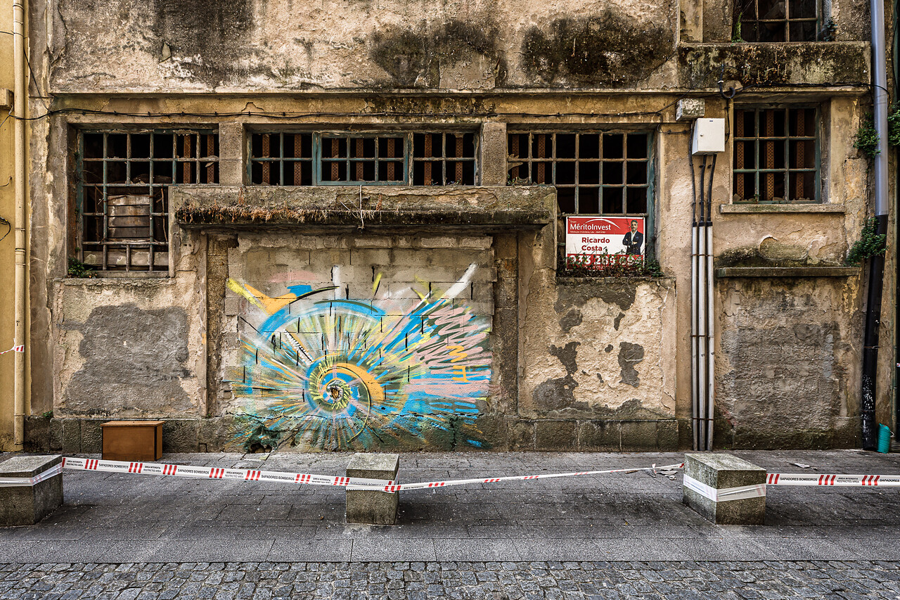 Ancienne façade délabrée à Vila Nova de Gaia, près de Porto, avec murs en pierre et enduit écaillé, fenêtres à petits carreaux aux vitres brisées, traces d’humidité et de végétation. Un graffiti coloré bleu, jaune et rose orne une ouverture murée au centre du bâtiment, tandis qu’une affiche de l’agence immobilière MéritoInvest apparaît dans une fenêtre à droite. Des rubans de sécurité barrent le trottoir au premier plan.