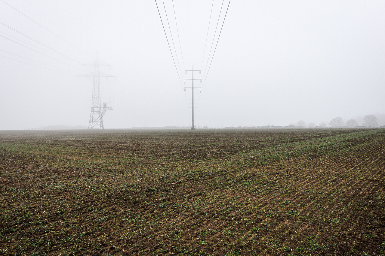 Un champ à Heisdorf avec des lignes électriques et des pylônes dans un paysage brumeux. Les lignes convergent vers un pylône central.