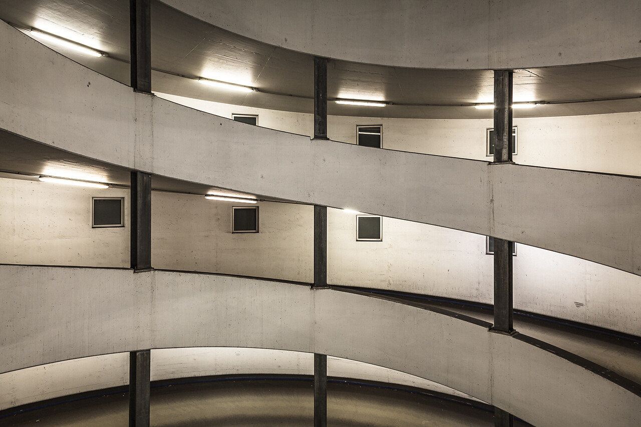 Parking en spirale avec des rampes en béton incurvées, des fenêtres carrées, des luminaires fluorescents et des piliers de soutien.