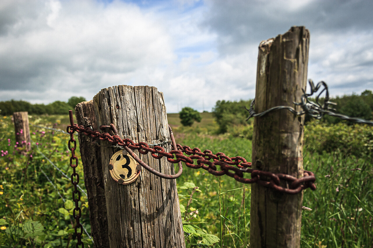 Clôture rustique en bois à Marloie en Belgique, avec une chaîne en métal rouillé et un médaillon estampillé du chiffre 3 fixé sur un poteau. Le paysage montre un champ avec des arbres et un ciel nuageux.