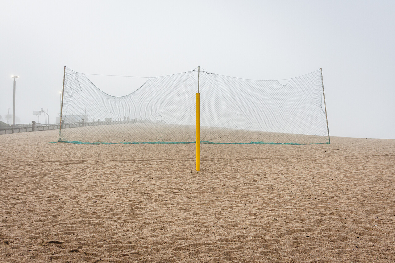 Plan large  et symétrique d'un terrain de beach-volley déserté, sous une lumière diffuse due au brouillard. Au centre, un filet vert séparant le terrain de la plage et un poteaux de couleur jaune à l'avant plan.
