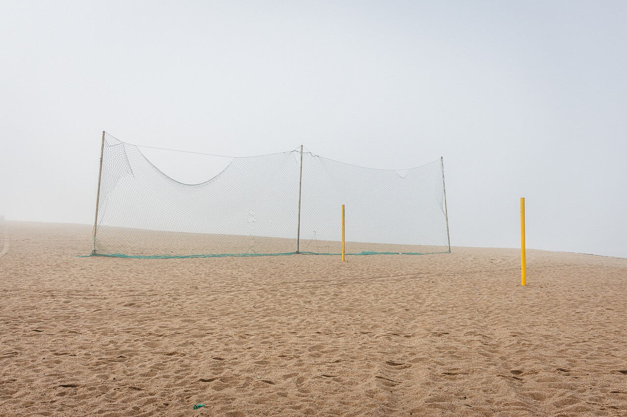 Plan large et minimaliste d'un terrain de beach-volley déserté, sous une lumière diffuse due au brouillard. Au centre, un filet vert est tendu pour séparer le terrain de la plage. Deux poteaux de couleur jaune, sans filet, se dressent à droite dans le sable.