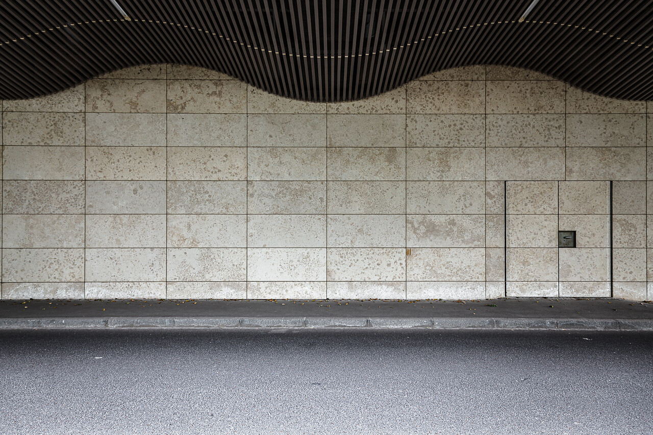 un pan de mur composé de carreaux rectangulaires avec un motif tacheté. Une petite porte est visible sur le côté droit du mur. Au-dessus du mur, on distingue une structure ondulée et sombre faite de lattes, qui fait partie du toit. Le sol est pavé d'asphalte, et une bordure sépare le mur de la route.