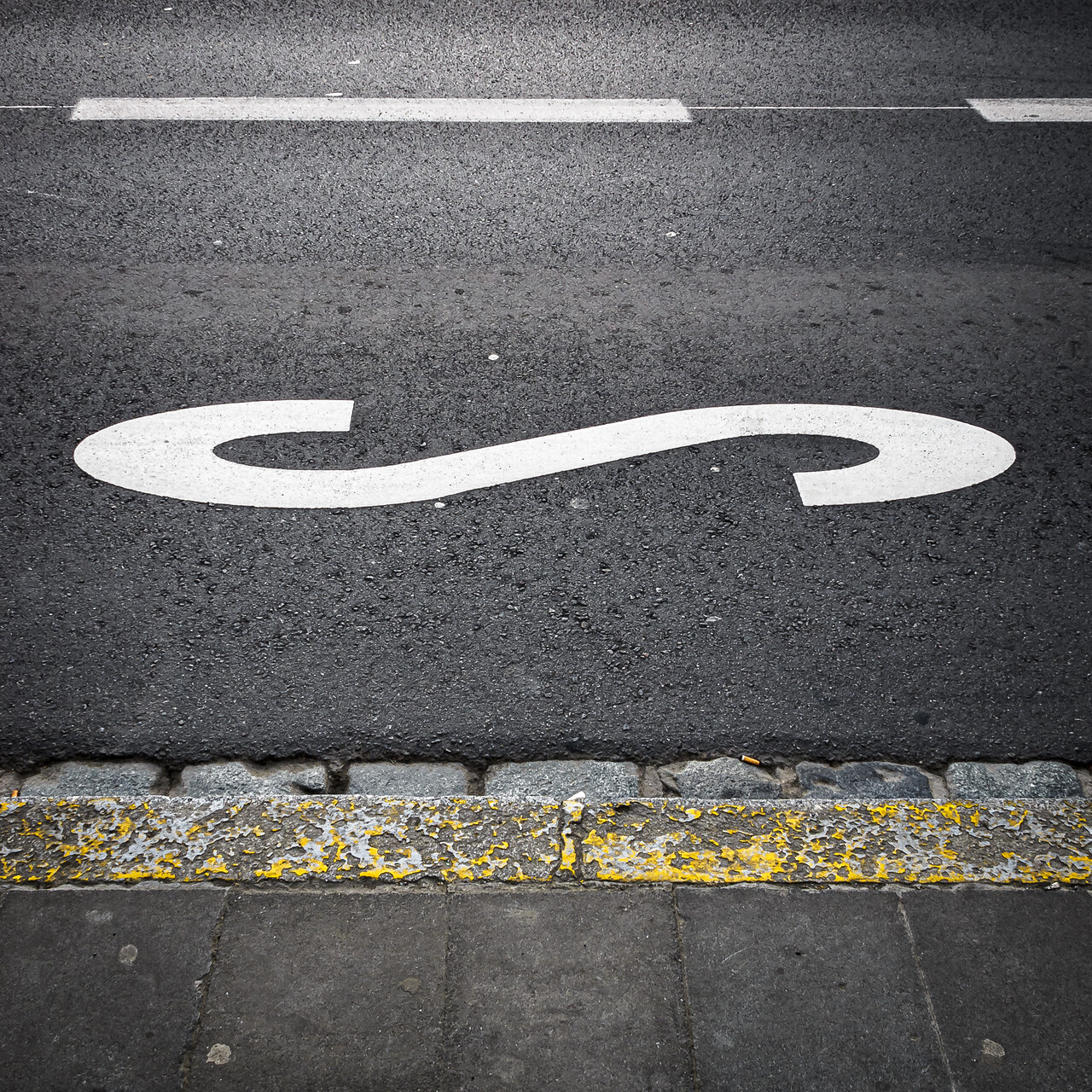 Un marquage routier en forme de 'S' blanc sur une route noire avec une ligne blanche discontinue en haut et un trottoir à la bordure jaune en bas.