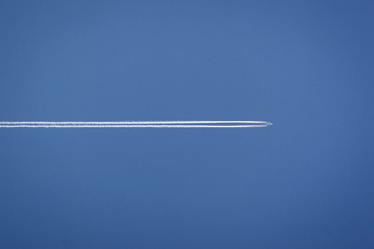 Traînée de condensation blanche laissée par un avion visible dans un ciel bleu uni de Sombreffe en Belgique. La traînée est horizontale et fine, s'étendant sur une grande partie de l'image.