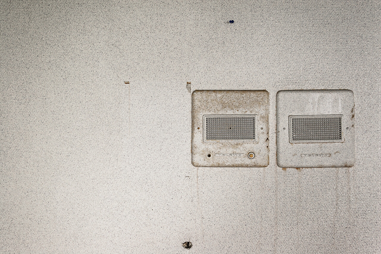 Deux grilles d'interphones de la marque Televic montées sur mur texturé, le tout sali et usé par le temps.