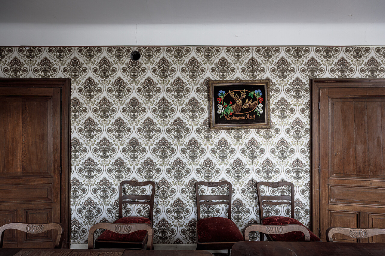 Austère salle à manger meublée de chaises aux coussins de velours rouge. Avec pour seule décoration un cadre illustré 'Waidmanns Heil!' ...