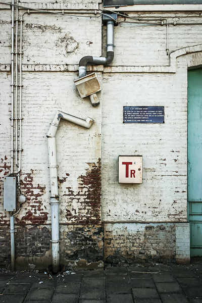 Un mur en briques de la gare de Gouy-lez-Piéton avec des tuyaux, une plaque bleue au lettrage blanc et une boîte en aluminium avec 'Tr' en rouge.