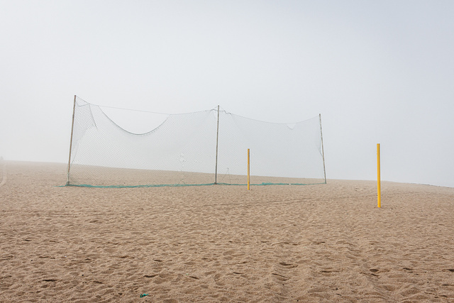 Plan large et minimaliste d'un terrain de beach-volley déserté, sous une lumière diffuse due au brouillard. Au centre, un filet vert est tendu pour séparer le terrain de la plage. Deux poteaux de couleur jaune, sans filet, se dressent à droite dans le sable.