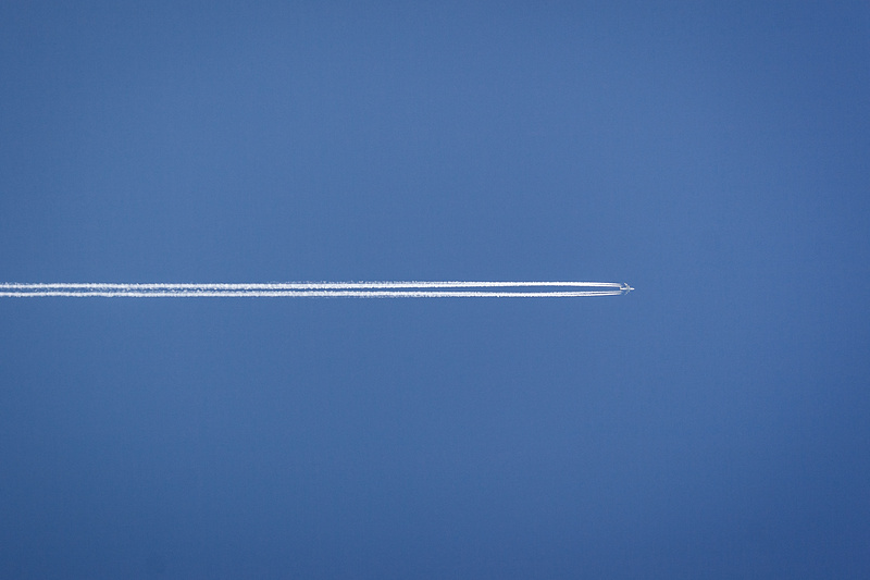 Traînée de condensation blanche laissée par un avion visible dans un ciel bleu uni de Sombreffe en Belgique. La traînée est horizontale et fine, s'étendant sur une grande partie de l'image.