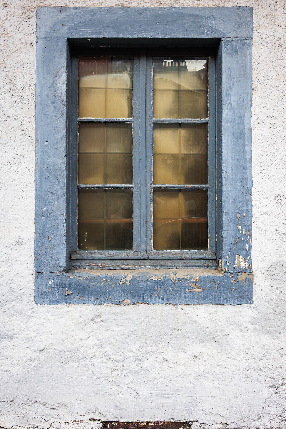 Plein cadre sur une fenêtre aux vitres jaunies et son encadrement peint en bleu sur un mur blanc.