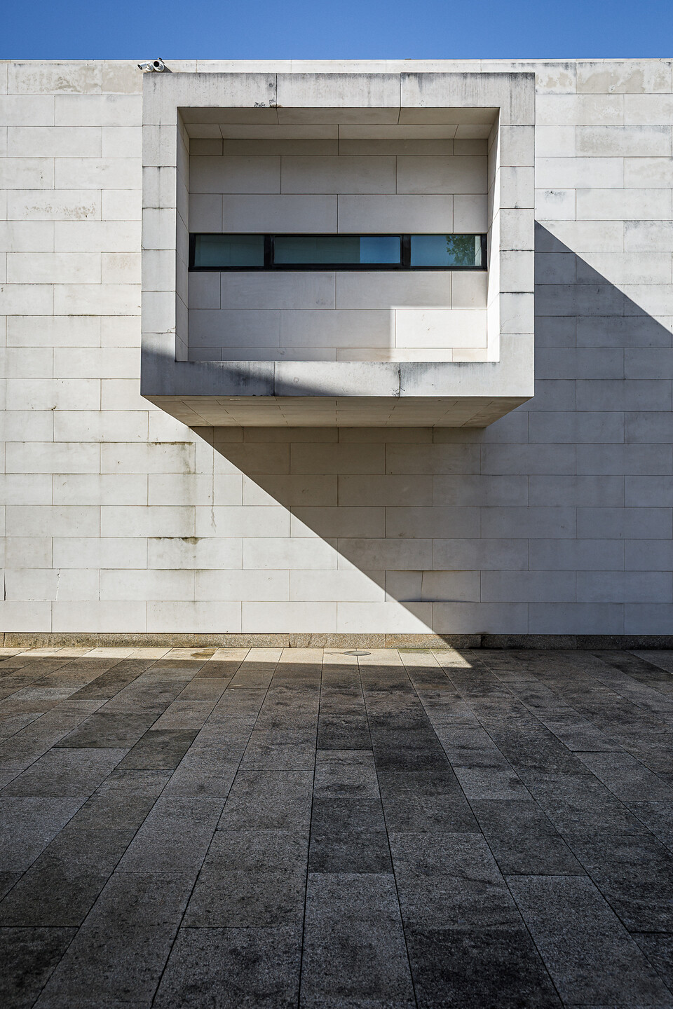 Façade en pierres calcaires blanches d'un bâtiment moderne (Centro de Memória de Vila do Conde, Porto, Portugal), avec un volume carré en saillie abritant une fenêtre horizontale. Une ombre triangulaire nette est projetée sous la saillie, accentuant le contraste avec le sol en dalles de granit foncé.