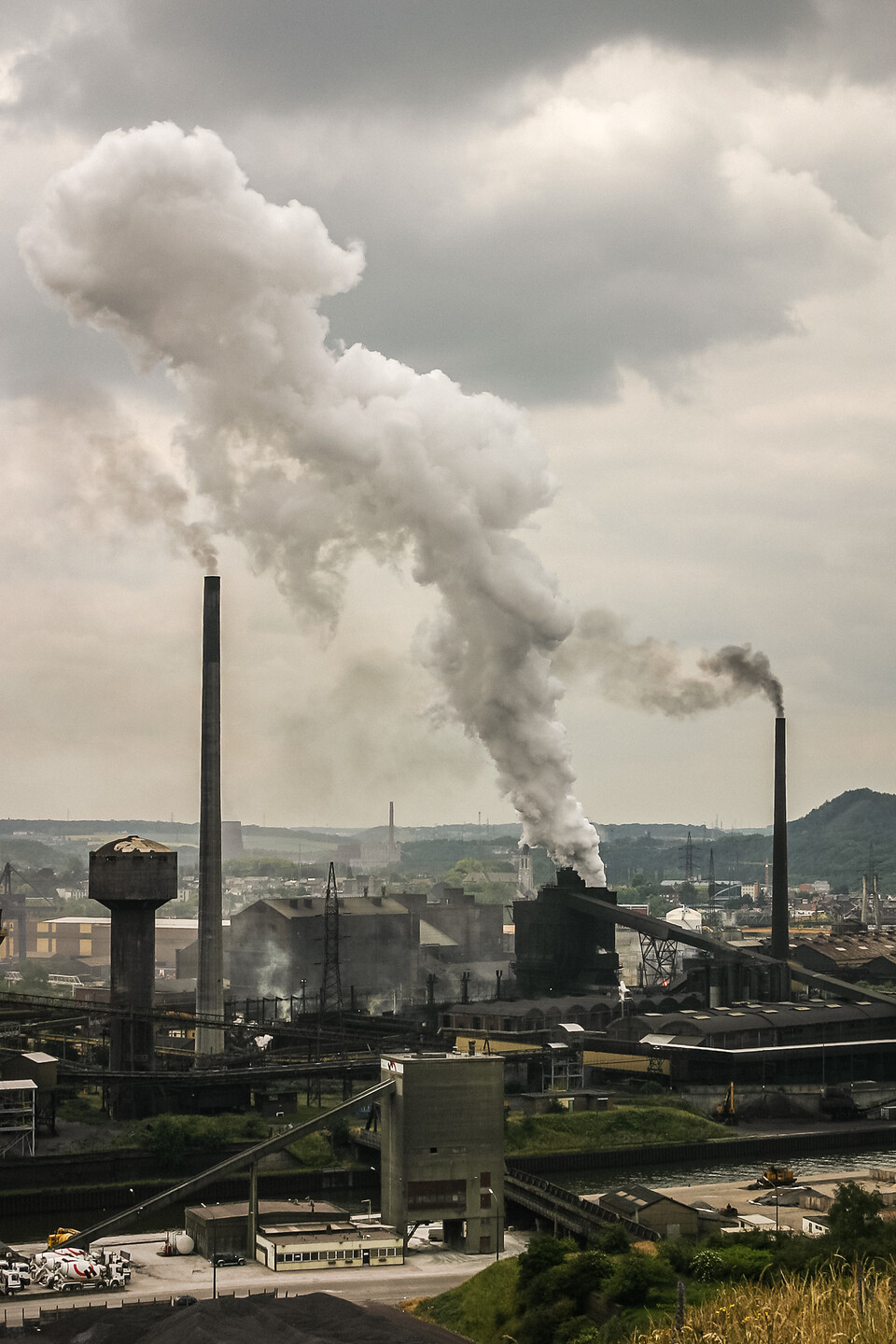 Paysage industriel à Charleroi en Belgique, avec une usine émettant de la fumée blanche dense, des cheminées noires, des bâtiments industriels et un ciel nuageux gris.