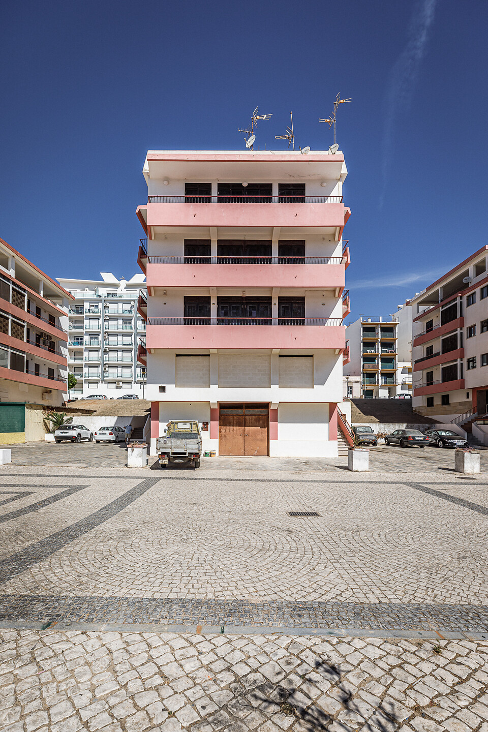 Immeuble résidentiel moderne de plusieurs étages aux façades blanches et roses, avec balcons, antennes paraboliques et véhicules stationnés devant, sur une place en calçada portuguesa.