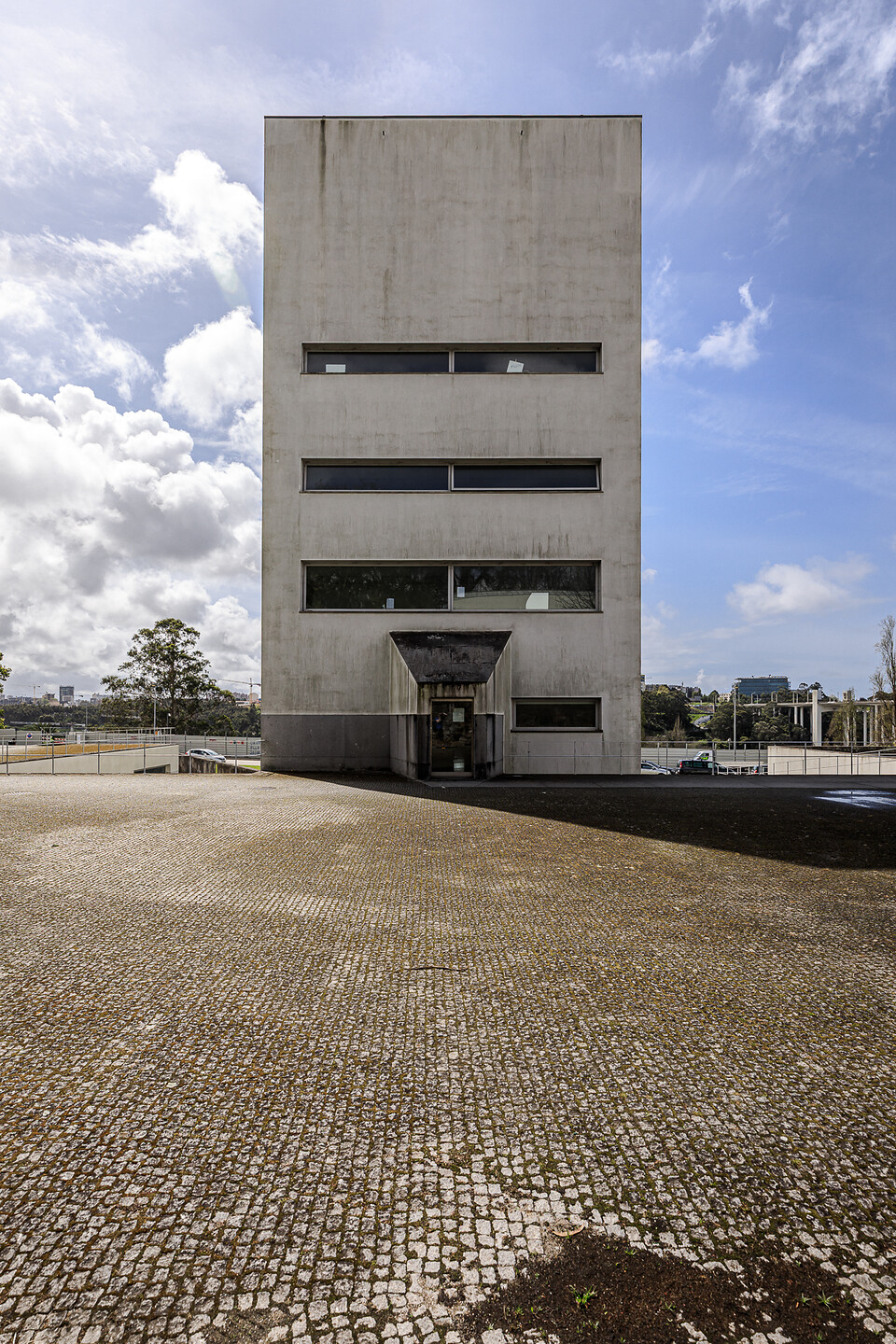 Façade bétonnée de la 'Torre H' avec entrée en avancée, vue depuis une esplanade pavée sous un ciel nuageux. (Massarelos, Porto, Portugal)