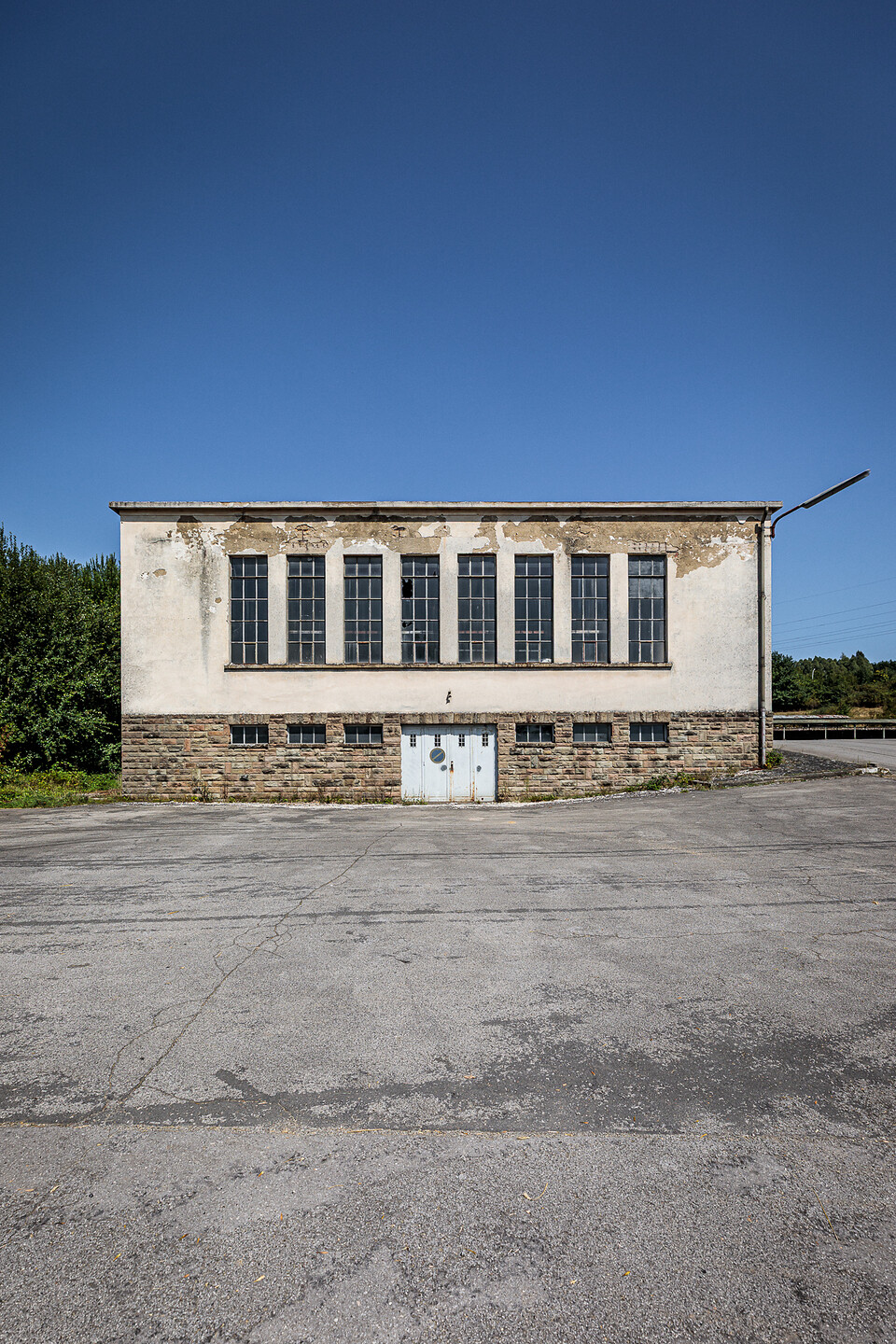 Vue frontale du bâtiment technique abandonné de l'ancienne laiterie Laduno-Luxlait, avec sa façade en pierre et en crépi défraîchi, ses grandes fenêtres verticales et sa cour asphaltée sous un ciel bleu.