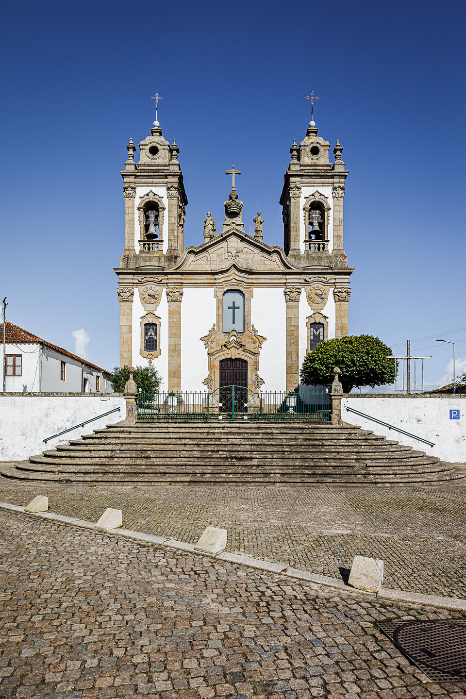 Église baroque avec deux clochers, façade ornée de statues, entrée centrale en bois surmontée d’une croix, située au sommet d’escaliers en pierre. Cour pavée, ciel bleu, arbres et bâtiment à gauche, panneau de stationnement à droite.