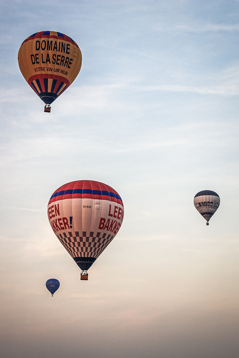 Quatre montgolfières de différentes tailles et couleurs flottant dans un ciel de Baisy-Thy en Belgique. La montgolfière principale est orange avec l'inscription 'DOMAINE DE LA SERRE', une autre est à damier gris et blanc avec l'inscription 'LEEN BAKKER!', et les deux autres sont plus petites et plus éloignées.