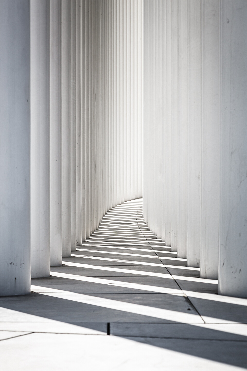 Perspective d'une allée de colonnes blanches de la Philharmonie de la ville de Luxembourg, avec les ombres projetées sur le sol en béton.
